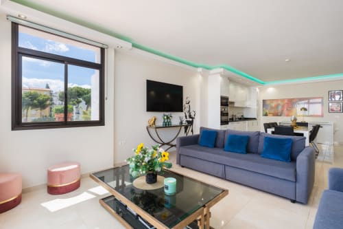 Modern Marbella apartment living room with gray sofa, blue pillows, large window with city views, and open kitchen layout
