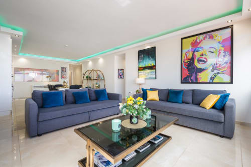 Modern Marbella apartment living room with blue sectional sofas, green neon ceiling, colorful pop art, and glass coffee table
