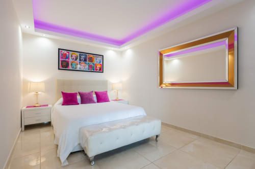 Modern bedroom at Marbella Home Rentals with purple LED ceiling lights, white bedding, magenta pillows, gold-framed mirror,