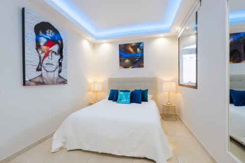 Modern bedroom in Marbella beachfront apartment with white bed, blue cushions, David Bowie artwork, LED ceiling lights, and