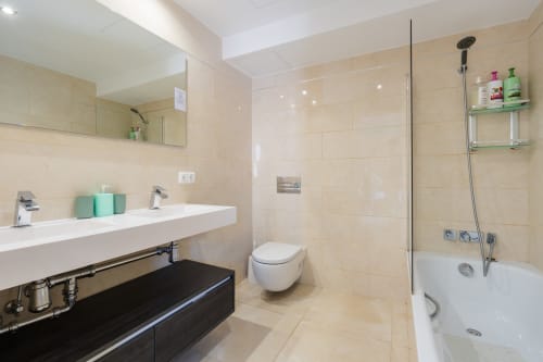 Modern luxury bathroom in Marbella beachfront apartment with cream tiles, dual sink, glass shower enclosure, and contemporary