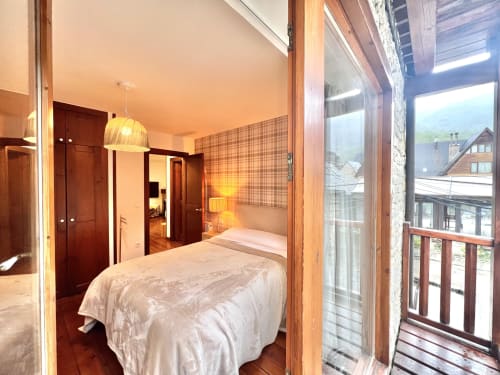 Dormitorio moderno en Vielha con cama doble, muebles de madera, vistas a las montañas desde el balcón e iluminación cálida.