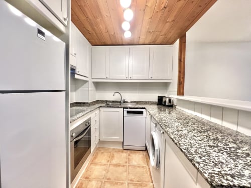 Cocina moderna en apartamento de Vielha con armarios blancos, encimeras de granito, electrodomésticos de acero inoxidable y techo de madera