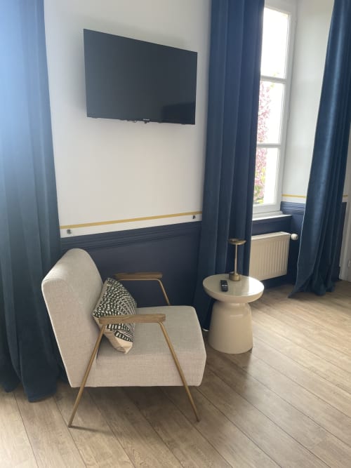 Chambre moderne au domaine de Marculfo avec fauteuil beige, TV murale et fenêtres donnant sur le jardin avec rideaux bleus