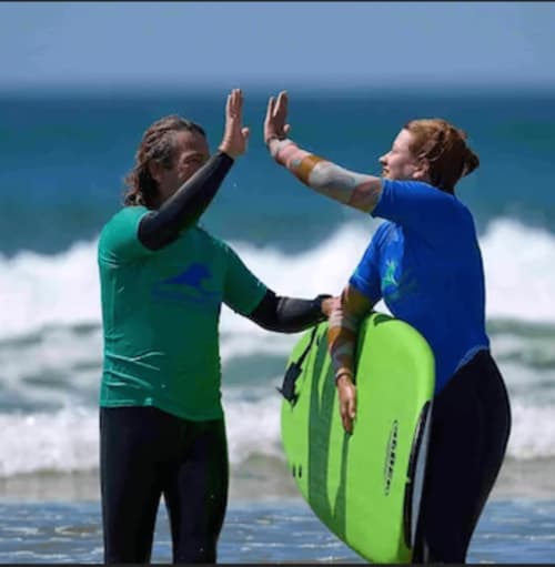 Duas mulheres trocando high-five na praia da Costa da Caparica, uma delas segurando uma prancha de surf verde