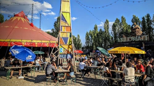 Zone extérieure du festival Killowatt avec chapiteau de cirque rouge, tour colorée, foule, parapluies et peupliers près d'Ivry-sur-Seine