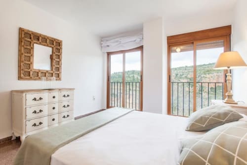 Dormitorio luminoso en Rustikalpuente con ropa de cama blanca, muebles de madera y vistas panorámicas al campo desde las puertas del balcón