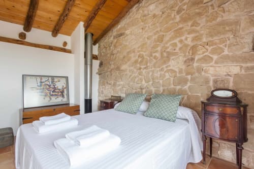 Dormitorio rústico con pared de piedra, techo de vigas de madera, cama blanca con cojines verdes y mobiliario de época