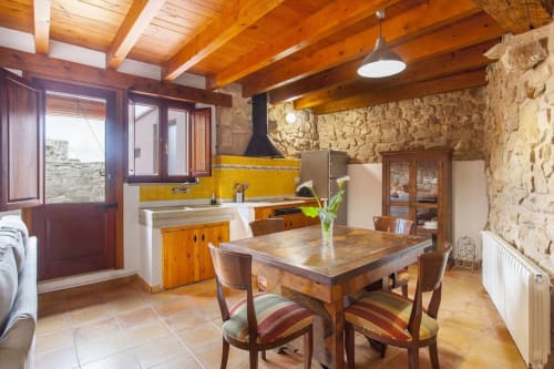 Cocina y comedor rústico con vigas de madera, muros de piedra, acentos amarillos y mobiliario tradicional en