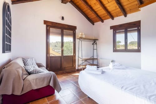 Dormitorio del apartamento Rustikalpuente con vigas de madera, azulejos de terracota, cama blanca, diván y vistas al paisaje rural