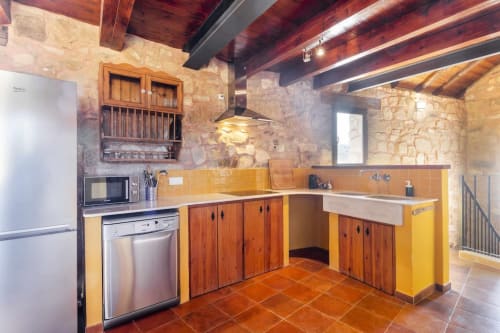 Cocina rústica moderna en apartamento Rustikalpuente con muebles de madera, detalles en azulejos amarillos, paredes de piedra y vigas expuestas.