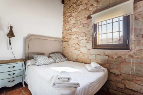 Dormitorio rústico en Rustikalpuente con muros de piedra vista, cama tapizada beige y ventana con vistas al campo español