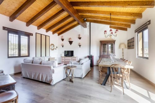 Apartamento Rustikalpu con vigas de madera vista, sofás blancos, mesa de comedor y decoración rústica en España
