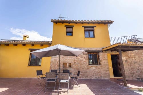 Patio del apartamento Rustikalpuente con edificio amarillo, muros de piedra, mesa de comedor bajo sombrilla y patio de terracota.