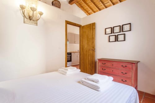 Dormitorio luminoso con cama blanca, cómoda roja y puerta de madera hacia la cocina en el apartamento Rustikalpuente de España