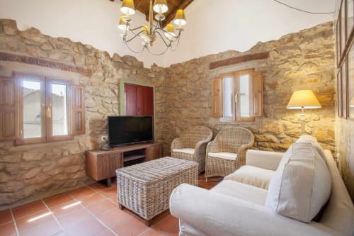 Sala de estar rústica de piedra con sofá beige, sillas de mimbre, televisor y piso de terracota