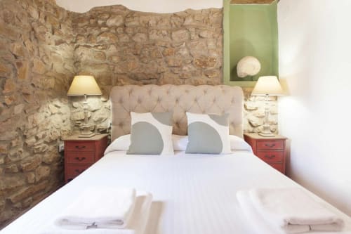 Dormitorio del apartamento Rustikalpuente con muros de piedra, cabecera capitonada beige, mesitas rojas y ropa de cama blanca.