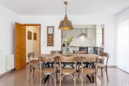 Comedor moderno con mesa de madera, sillas de ratán, lámpara colgante de latón y cocina abierta en Rustikalpuente, España