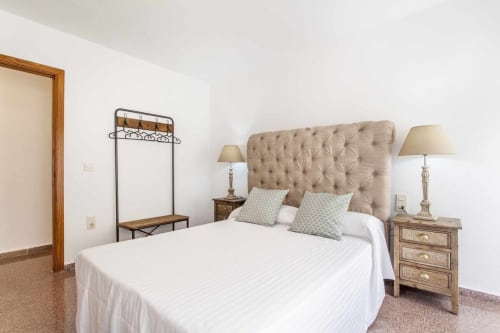 Dormitorio del apartamento Rustikalpuente con cabecero tapizado beige, ropa de cama blanca, mesitas de madera y perchero metálico