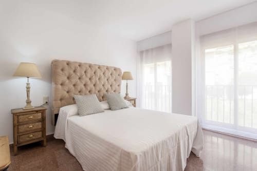 Dormitorio luminoso en apartamento Rustikalpuente con cabecero capitoné, ropa de cama blanca, mesillas de madera y puertas de vidrio