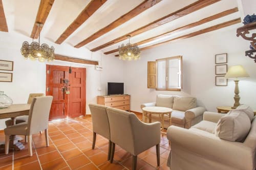 Sala de estar rústica con vigas de madera vista, suelo de baldosas de terracota, muebles crema y puerta de entrada roja