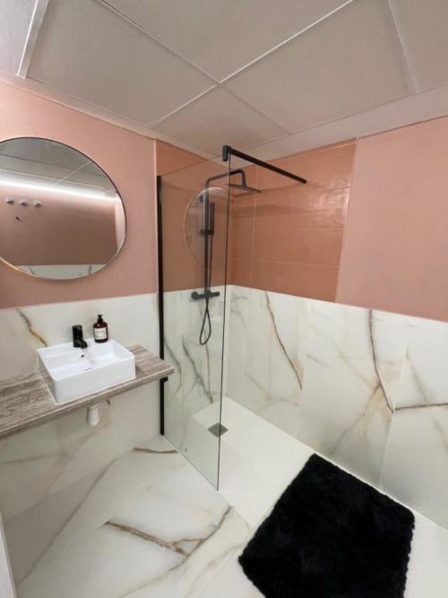 Baño moderno en Agata 10 Apartamentos, Alicante con paredes rosa, mampara de ducha en mármol, espejo redondo y lavabo blanco.