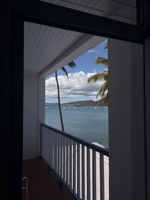 Vue de balcon de la chambre de l'hôtel La Dunette à Sainte-Anne avec palmier, bateaux amarrés et montagnes côtières sous ciel bleu