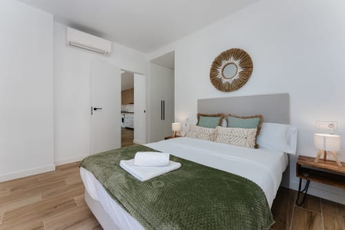 Dormitorio moderno en Ab Be Apartments, Fuengirola con cama, manta verde, espejo redondo y acceso a balcón con vistas al mar