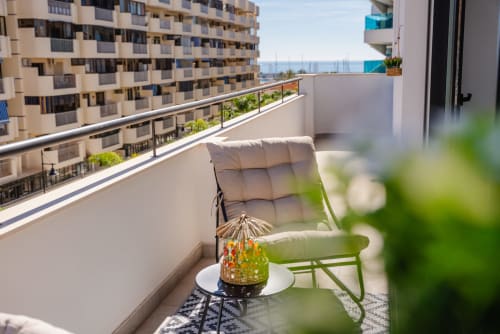 Balcón soleado en Ab Be Apartments de Málaga con silla acolchada, flores en maceta y vistas al mar Mediterráneo.