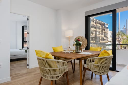 Moderna zona de comedor en apartamento aMar Fuengirola con mesa de madera, sillas tejidas, cojines amarillos y balcón con vistas al mar