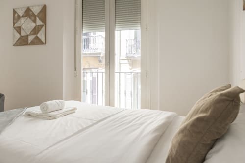 Dormitorio luminoso en Ab Be Apartments Málaga con ropa de cama blanca, puertas al balcón con vistas a la ciudad, arte geométrico en las paredes