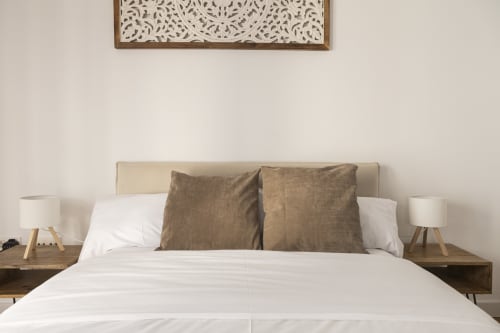 Moderno dormitorio en Ab Be Apartments de Málaga con cabecero beige, ropa de cama blanca, cojines marrones y mesillas de madera