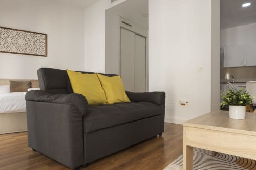 Sala de estar de apartamento estudio moderno con sofá oscuro, almohadas amarillas, mesa de madera y kitchenette en Ab Be Apartments