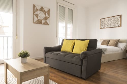 Moderno apartamento estudio en Málaga con sofá oscuro, almohadas amarillas, mesa de madera y cama con vistas a la ciudad