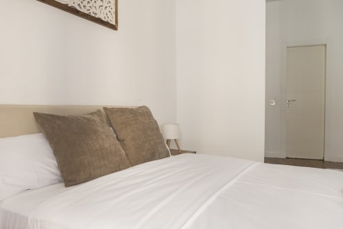 Dormitorio moderno en Ab Be Apartments, Málaga, con ropa de cama blanca, almohadas beige, lámpara de noche y puerta al baño