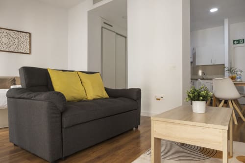 Zona de estar de estudio moderno con sofá oscuro, cojines amarillos, mesa de madera y cocina americana en Ab Be Apartments, Málaga