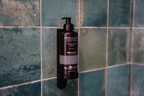 Botella dispensadora oscura de jabón líquido para manos sobre pared de azulejos verdeazulados en baño de apartamentos ABBE en Málaga