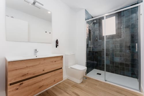 Baño moderno en Ab Be Apartments, Málaga con vanitory de madera, lavamanos blanco, inodoro y ducha abierta con muros de azulejos oscuros