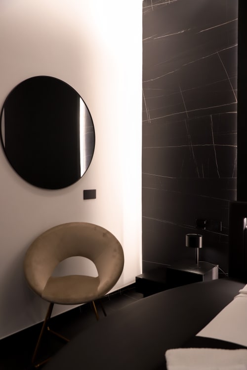 Bagno moderno con poltrona curva beige, specchio tondo nero e parete in piastrelle scure presso Privitera Rooms a Grammichele