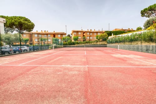 Red clay tennis court with white lines at Finques Garcia Enrich apartment in Palafrugell, surrounded by green fencing and