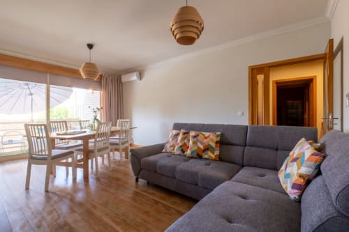 Sala de estar moderna com sofá cinza, mesa de jantar em madeira, luminárias pendentes e varanda com vista para a cidade no apartamento EcoHost