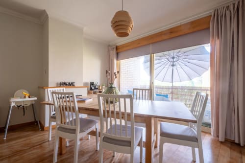 Sala de jantar luminosa com mesa de madeira, cadeiras brancas, luminária pendente e vista para a varanda no apartamento EcoHost, Portugal