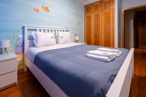 Quarto luminoso no apartamento EcoHost FamilyHub com paredes temáticas oceânicas, cama branca e azul, guarda-roupa em madeira e piso de madeira