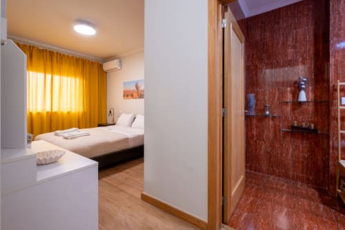 Quarto moderno com banheiro integrado no apartamento EcoHost, com cama, cortinas douradas e área de chuveiro em mármore vermelho