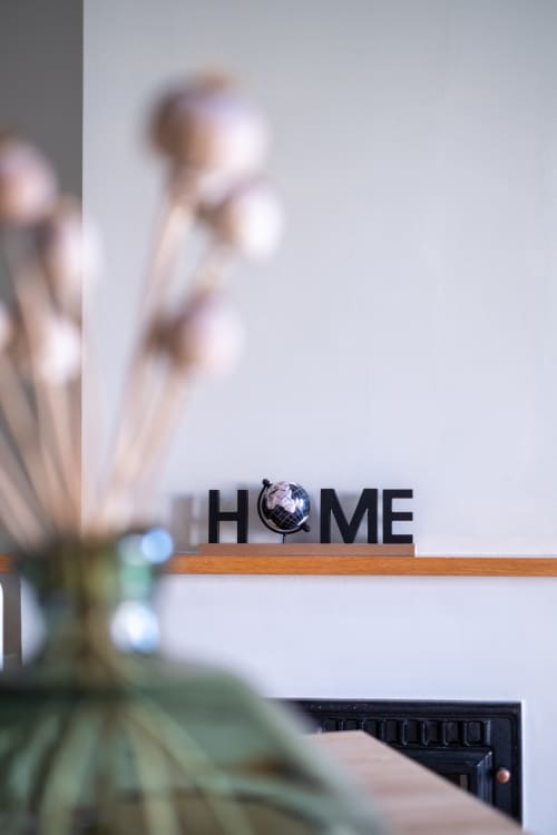 Placa decorativa HOME em prateleira de madeira com flores desfocadas e parede branca ao fundo no apartamento EcoHost