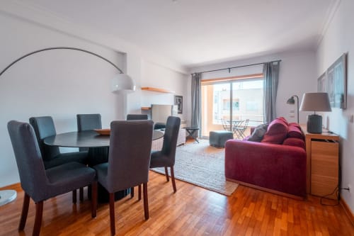 Sala de estar moderna no apartamento OceanGreen da EcoHost com sofá bordô, mesa de jantar, pisos de madeira e vista para o oceano