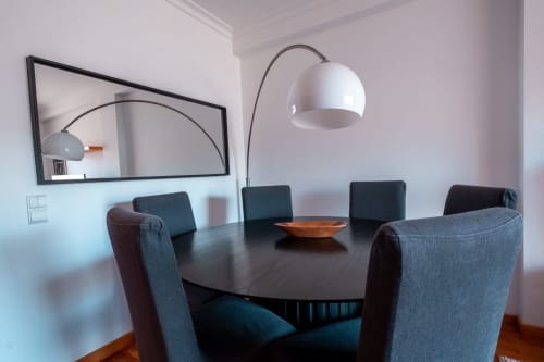 Sala de jantar moderna no EcoHost com mesa redonda preta, cadeiras estofadas cinzentas, luminária arco branca e espelho retangular na parede