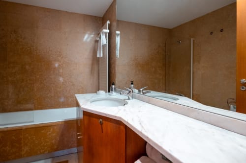 Banheiro moderno em apartamento EcoHost com bancada de mármore, azulejos marrom, pia, espelho e banheira com vidro