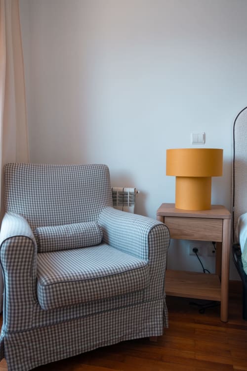 Canto aconchegante do quarto na EcoHost com poltrona xadrez, criado de madeira e luminária amarelo-mostarda em apartamento com vista para o jardim
