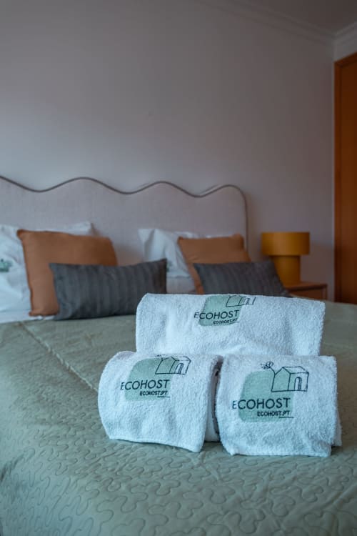 Toalhas brancas com marca EcoHost dispostas em cama do apartamento OceanGreen, com cabeceira bege visível.